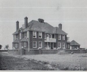 A two-story brick building with multiple chimneys and a balcony, enclosed by a low wall, viewed from a gravel foreground.