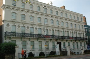 A large, pale, four-story Victorian terrace building with decorative relief carvings and a black wrought-iron balcony.