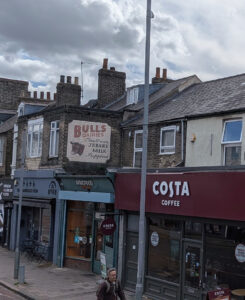 A brick building on Hills Road features a faded painted advertisement for Bulls Dairies above a row of modern shopfronts.