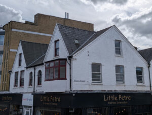 White brick building with a dark slate roof and a red-framed bay window, featuring a Little Petra restaurant on the ground.