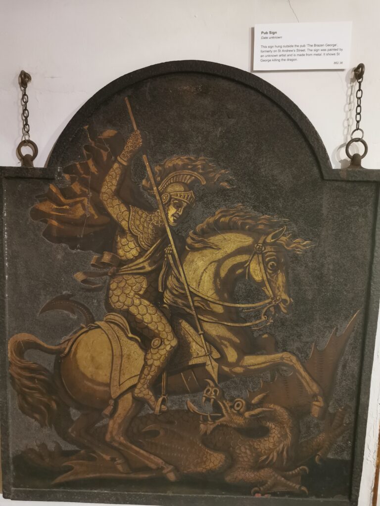 A metal pub sign shows a golden armored knight on horseback brandishing a long spear over a dragon on a dark background.