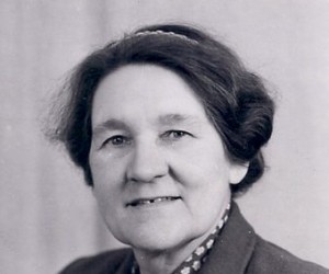 A black and white head-and-shoulders portrait of a middle-aged woman with dark wavy hair wearing a collared patterned blouse.