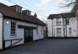 Two white painted brick buildings meet at a corner, separated by a grey paved courtyard and featuring dark window frames.