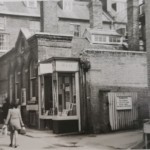 A brick building in Post Office Terrace with a shop corner marked R&M, beside a corrugated fence with a danger warning sign.