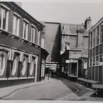 A narrow street between a dark brick building with oval windows and a modern glass-fronted office building in Cambridge.