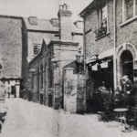 Narrow cobblestone alleyway in 1873 flanked by brick buildings with arched doorways, a lantern, and two figures near a table.