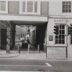 A narrow alleyway sits between a shop entrance and a Barclays Bank, with a small car parked inside guarded by stone bollards.