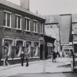 Pedestrians walk along a narrow street lined with brick buildings and shops with oval windows featuring sale advertisements.