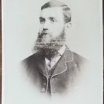 A studio portrait of a man with a thick beard and short dark hair wearing a buttoned jacket and a high collared shirt.