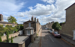 A street scene in Cambridge with yellow brick terraced houses, a paved driveway, parked cars, and a blue sky with clouds.