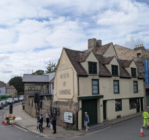 The Museum of Cambridge, a cream-colored building with dark green trim, stands on a street corner with pedestrians nearby.