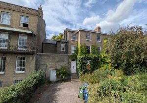 Yellow brick terrace houses with sash windows, a white front door, and ivy-covered walls overlooking a gravel driveway.