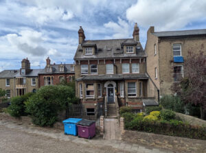 A three-story yellow brick house with a central entrance and two blue and purple wheelie bins on the pavement in front.