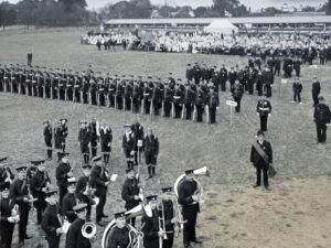 A large group of uniformed men in formation on a field, with a brass band in the foreground and spectators in the distance.