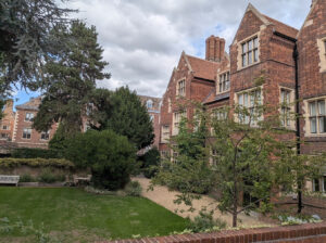 Red brick buildings with gabled roofs overlook a green lawn, a gravel path, and trees under a cloudy sky.