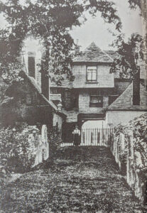 A path leads to a gated entrance between two historic buildings, with a person standing in the gateway under overhanging trees.