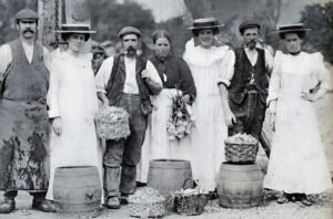 Seven people stand in a row behind barrels and baskets, wearing early twentieth-century work clothes and formal white dresses.