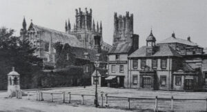 Black and white view of Ely Market Place with a low fence in the foreground, stone buildings, and the cathedral in the distance.