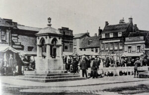 A stone fountain stands in a market square surrounded by stalls and people in period clothing in front of brick buildings.