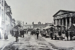 Black and white view of Ely Market Place with stalls, a horse-drawn carriage, and people walking past a classical building.