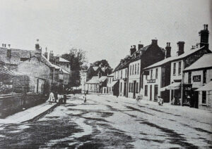 A black and white print of a street scene in Ely with terraced buildings, a few pedestrians, and a bicycle on the pavement.