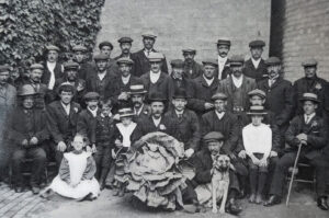 A large group of men and two children pose outdoors, with a giant cabbage placed prominently in the center foreground.
