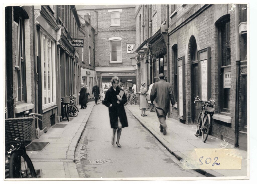 Pedestrians walk along a narrow brick-walled street in Cambridge, with bicycles parked against the buildings on both sides.